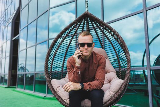 A cheerful young man in a shirt with a phone sits on the terrace of an office building after work, uses wireless internet, a positive male employee in glasses is talking on the phone