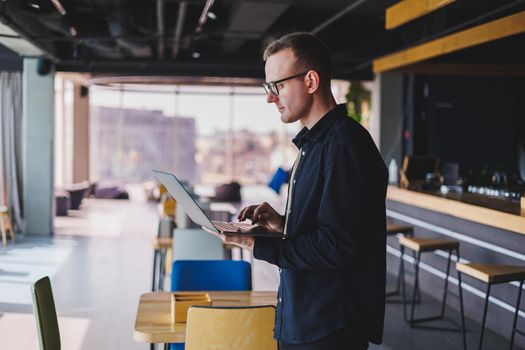 Male entrepreneur in black shirt and glasses standing in office with laptop in hand, successful corporate boss feeling good from rich lifestyle