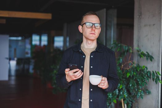 Portrait of a happy successful man in optical glasses holding a modern mobile phone and coffee in his hands, a cheerful blogger with a digital smartphone posing in a cafe