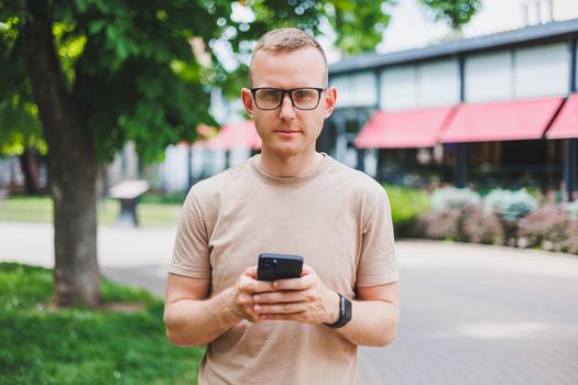 Portrait of handsome mature businessman holding messenger bag, wearing stylish eyeglasses looking at camera. Smiling middle aged man using smartphone, standing on the street. Mobile banking concept
