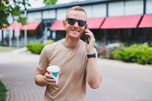 A bearded, smiling, stylish man in white shirt and sunglasses standing on the streets of the city and calling on mobile phone.