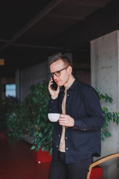 Portrait of a happy successful man in optical glasses holding a modern mobile phone and coffee in his hands, a cheerful blogger with a digital smartphone posing in a cafe