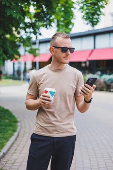 A bearded, smiling, stylish man in white shirt and sunglasses standing on the streets of the city and calling on mobile phone.