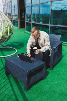 Confident young businessman, dressed in formal wear and stylish glasses, calls on a smartphone while sitting with coffee, on the terrace of a modern office building with a glass wall