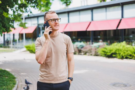 Portrait of handsome mature businessman holding messenger bag, wearing stylish eyeglasses looking at camera. Smiling middle aged man using smartphone, standing on the street. Mobile banking concept