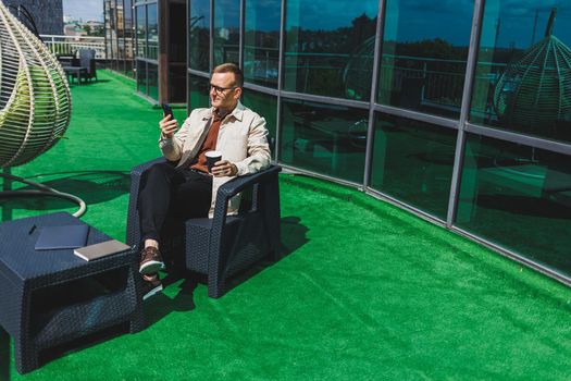 Confident young businessman, dressed in formal wear and stylish glasses, calls on a smartphone while sitting with coffee, on the terrace of a modern office building with a glass wall
