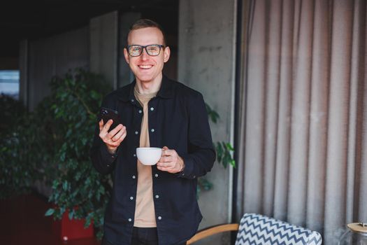 Portrait of a happy successful man in optical glasses holding a modern mobile phone and coffee in his hands, a cheerful blogger with a digital smartphone posing in a cafe