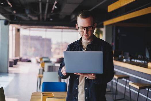 Male entrepreneur in black shirt and glasses standing in office with laptop in hand, successful corporate boss feeling good from rich lifestyle