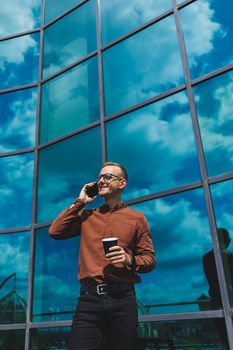 Attractive fashionable male businessman in a stylish shirt and trousers talking on the phone near the office building and drinking coffee, modern glass office