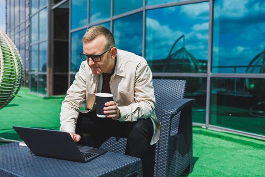 Confident young businessman, dressed in formal wear and stylish glasses, calls on a smartphone while sitting with coffee, on the terrace of a modern office building with a glass wall