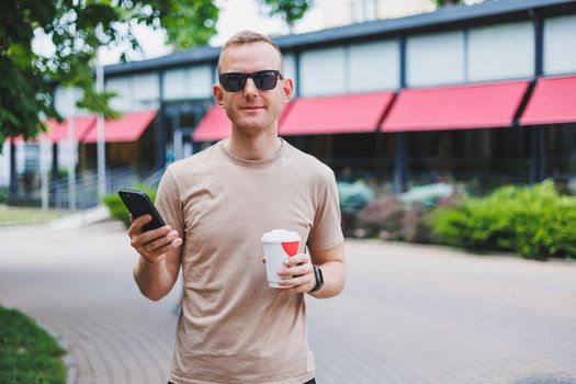 A bearded, smiling, stylish man in white shirt and sunglasses standing on the streets of the city and calling on mobile phone.