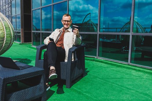 Confident young businessman, dressed in formal wear and stylish glasses, calls on a smartphone while sitting with coffee, on the terrace of a modern office building with a glass wall