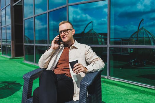 Confident young businessman, dressed in formal wear and stylish glasses, calls on a smartphone while sitting with coffee, on the terrace of a modern office building with a glass wall