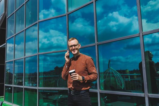 Attractive fashionable male businessman in a stylish shirt and trousers talking on the phone near the office building and drinking coffee, modern glass office