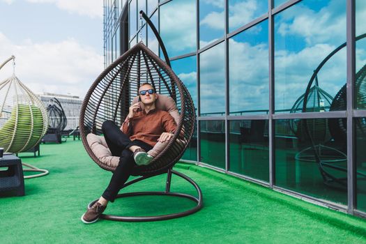 A cheerful young man in a shirt with a phone sits on the terrace of an office building after work, uses wireless internet, a positive male employee in glasses is talking on the phone