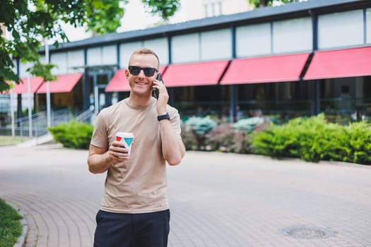 A bearded, smiling, stylish man in white shirt and sunglasses standing on the streets of the city and calling on mobile phone.