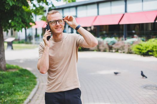 Portrait of handsome mature businessman holding messenger bag, wearing stylish eyeglasses looking at camera. Smiling middle aged man using smartphone, standing on the street. Mobile banking concept