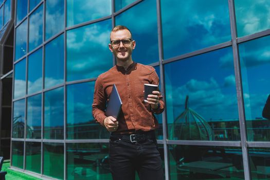 A young handsome attentive man in glasses with a laptop and a glass of coffee to go, stands on the terrace of an office. young smart developer