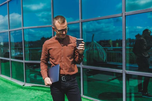 A young handsome attentive man in glasses with a laptop and a glass of coffee to go, stands on the terrace of an office. young smart developer