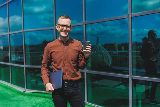 A young handsome attentive man in glasses with a laptop and a glass of coffee to go, stands on the terrace of an office. young smart developer