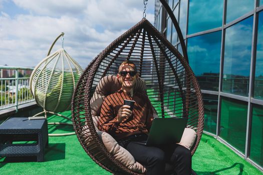 A businessman in formal wear sits on an armchair and looks at a laptop connected to a 4g wireless network on the terrace of a modern office while working remotely