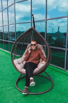 A cheerful young man in a shirt with a phone sits on the terrace of an office building after work, uses wireless internet, a positive male employee in glasses is talking on the phone