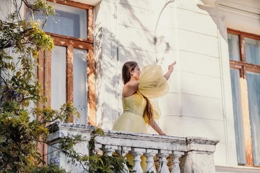 a beautiful smiling and kind woman in a gorgeous yellow dress stands on the balcony of an old vintage house.