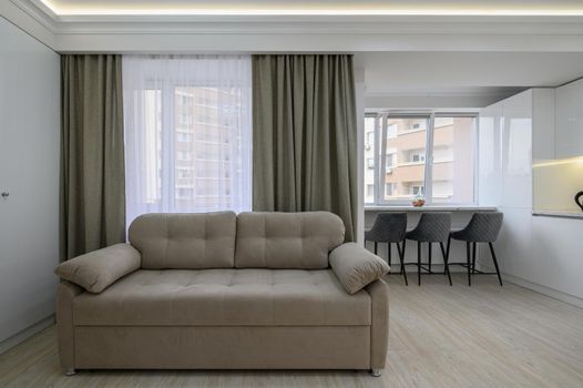 Living room with a couch and a kitchen in the background with large window an lights on the ceiling in studio apartment interior