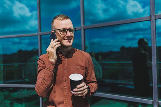 Attractive fashionable male businessman in a stylish shirt and trousers talking on the phone near the office building and drinking coffee, modern glass office