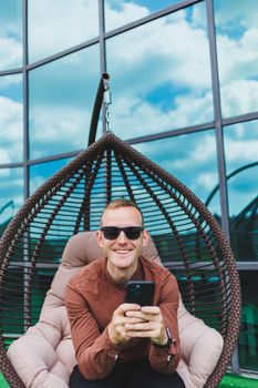 A cheerful young man in a shirt with a phone sits on the terrace of an office building after work, uses wireless internet, a positive male employee in glasses is talking on the phone