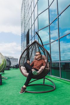 A cheerful young man in a shirt with a phone sits on the terrace of an office building after work, uses wireless internet, a positive male employee in glasses is talking on the phone