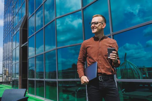 A young handsome attentive man in glasses with a laptop and a glass of coffee to go, stands on the terrace of an office. young smart developer