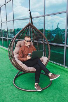 A businessman in formal wear sits on an armchair and looks at a laptop connected to a 4g wireless network on the terrace of a modern office while working remotely