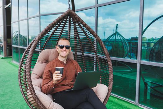 A businessman in formal wear sits on an armchair and looks at a laptop connected to a 4g wireless network on the terrace of a modern office while working remotely