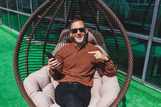 A cheerful young man in a shirt with a phone sits on the terrace of an office building after work, uses wireless internet, a positive male employee in glasses is talking on the phone