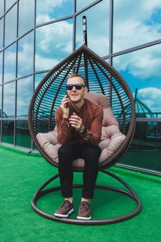 A cheerful young man in a shirt with a phone sits on the terrace of an office building after work, uses wireless internet, a positive male employee in glasses is talking on the phone