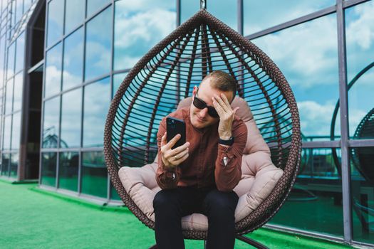 A cheerful young man in a shirt with a phone sits on the terrace of an office building after work, uses wireless internet, a positive male employee in glasses is talking on the phone