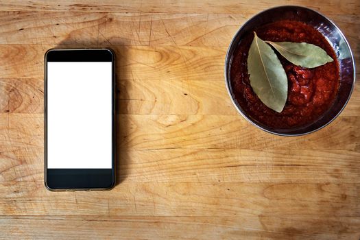 old wooden board with tomato sauce and laurel leaves in a bowl, with a smart-phone. Flat lay, top view with copy space