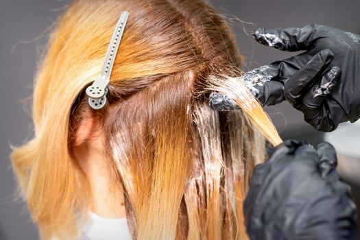 Hands of the hairdresser are applying the dye with gloved fingers on the red hair of a young red-haired woman in a hairdresser salon, close up
