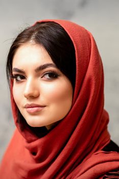The fashionable young woman. Portrait of the beautiful female model with long hair and makeup in a red scarf. Beauty young caucasian woman on the background of a gray wall