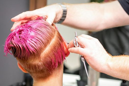 Haircut of dyed short pink wet hair of young caucasian woman by a male hairdresser in a barbershop