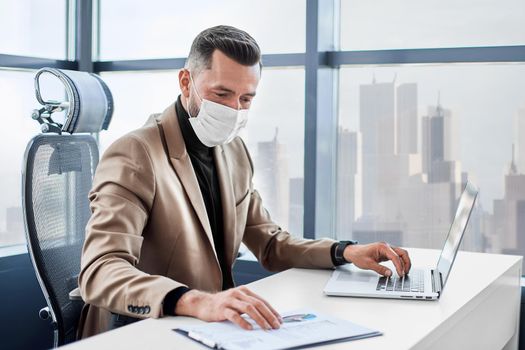 businessman in a protective mask working on a laptop in the office. side view.