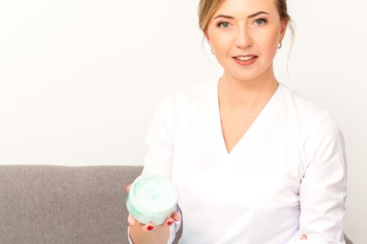 Close-up portrait of young smiling female caucasian healthcare worker standing with jar of cream beauty product on white background.