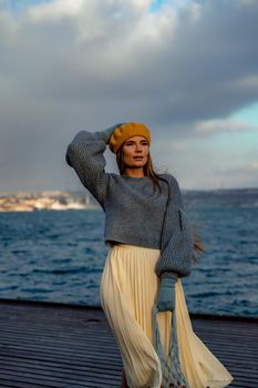 Outdoors fashion portrait of a beautiful middle aged woman walking on the beach. Marine background. Dressed in a stylish warm blue sweater, yellow skirt and beret