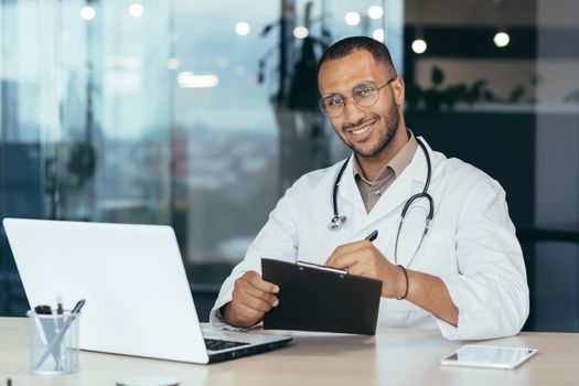 Portrait of successful male doctor, hispanic smiling and looking at camera, doctor in medical gown working inside modern clinic office, using laptop for work