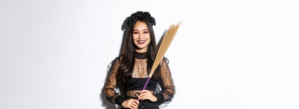 Portrait of smiling beautiful asian woman in witch costume holding broom and looking happy at camera, celebrating halloween, enjoying trick or treating, white background.