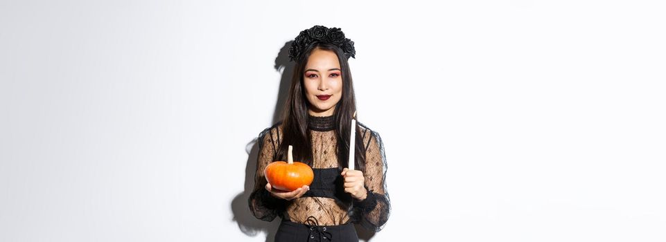Image of devious smiling asian witch in gothic dress, holding candle with pumpkin and looking at camera cunning, standing over white background.
