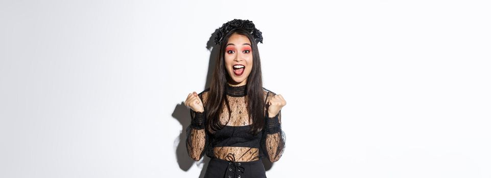 Image of happy asian gothic girl in halloween party dress, rejoicing over victory, triumphing and saying yes, standing over white background celebrating victory.
