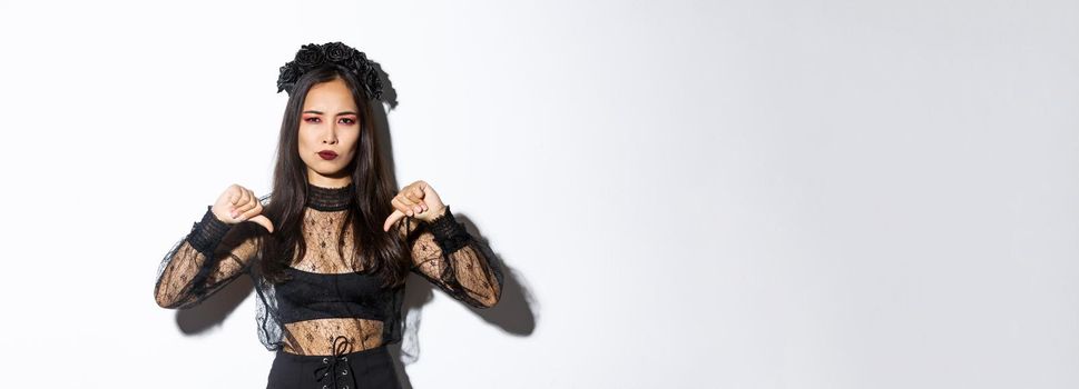 Image of disappointed asian woman in halloween dress of gothic undead girl showing thumbs-down, dislike and disagree with something bad, standing over white background.