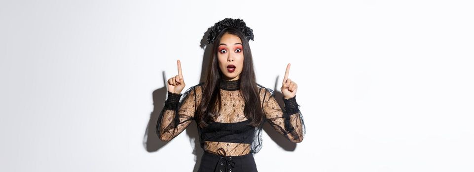 Impressed asian girl in black lace dress and wreat turn your attention on halloween promo, making announcement, pointing fingers up, showing banner, standing over white background.